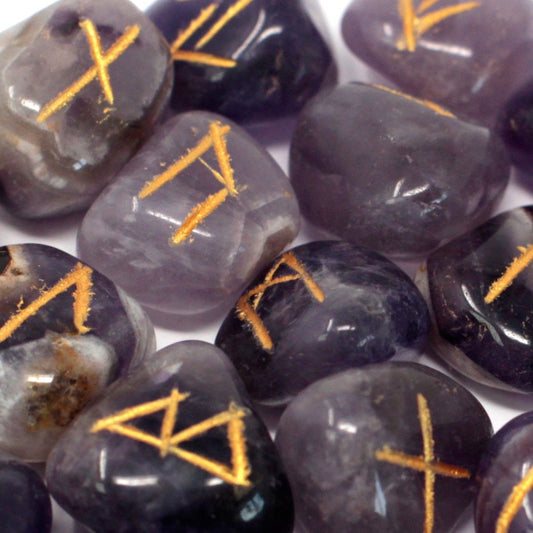 Tumbled amethyst stones engraved with traditional rune symbols laid out next to a small black drawstring pouch.