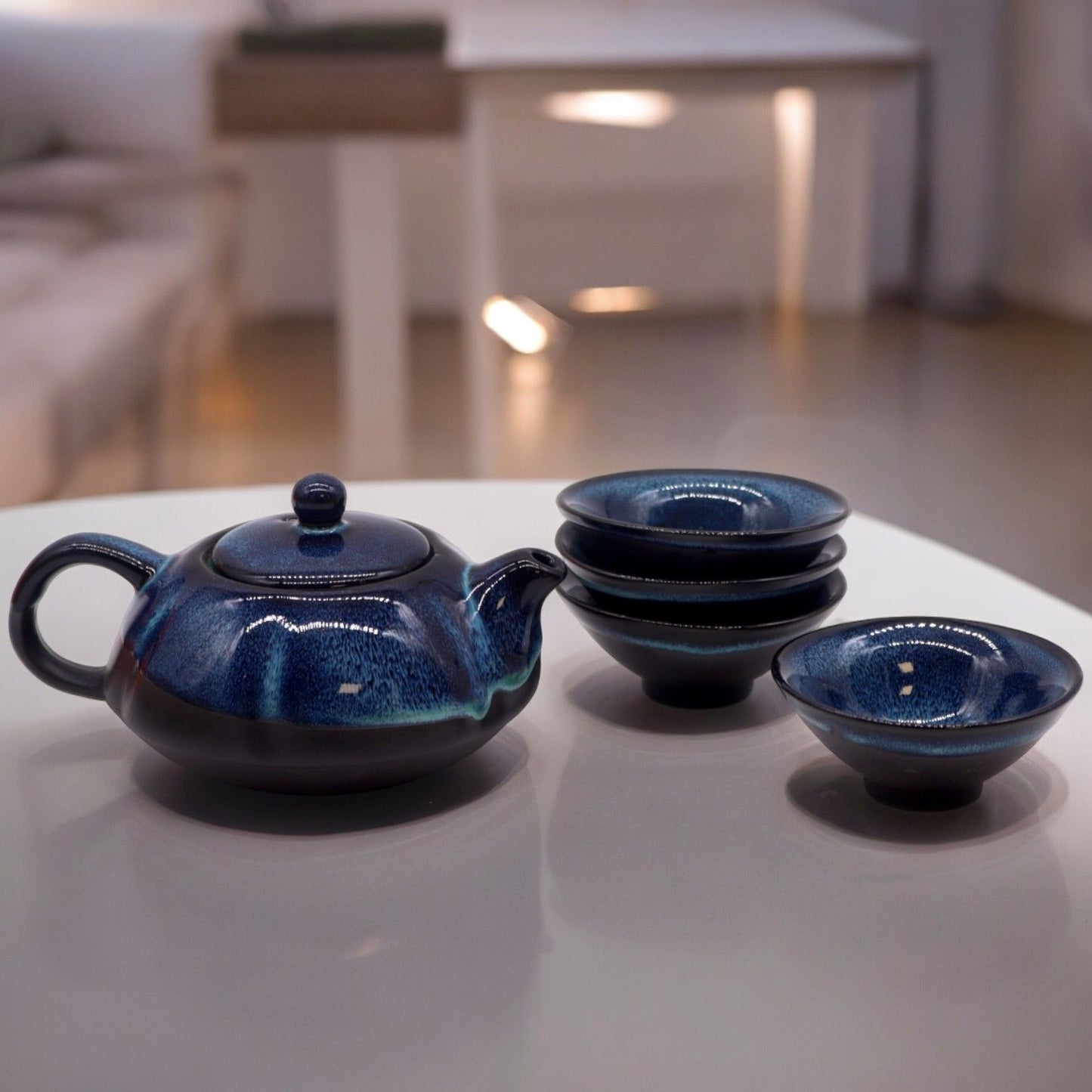 Elegant glossy blue ceramic teapot with subtle artistic gradients and four matching tea cups on a clean background.
