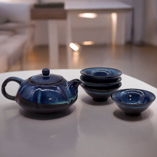 Elegant glossy blue ceramic teapot with subtle artistic gradients and four matching tea cups on a clean background.