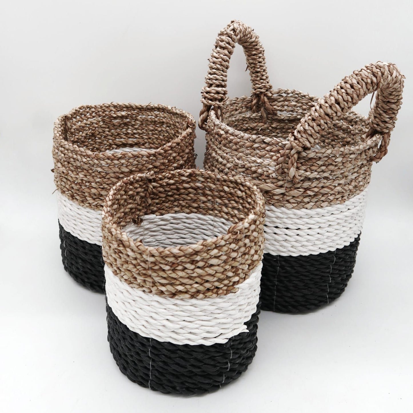 Set of three handwoven round seagrass baskets in dark grey, white, and natural tones with integrated handles.