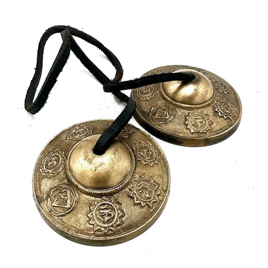 Pair of ornate Tibetan Tingsha bells joined by a leather strap, featuring embossed sacred Seven Chakra symbols.