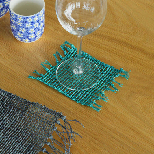 Set of four turquoise handwoven seagrass coasters featuring a natural fringe edge on a table setting.
