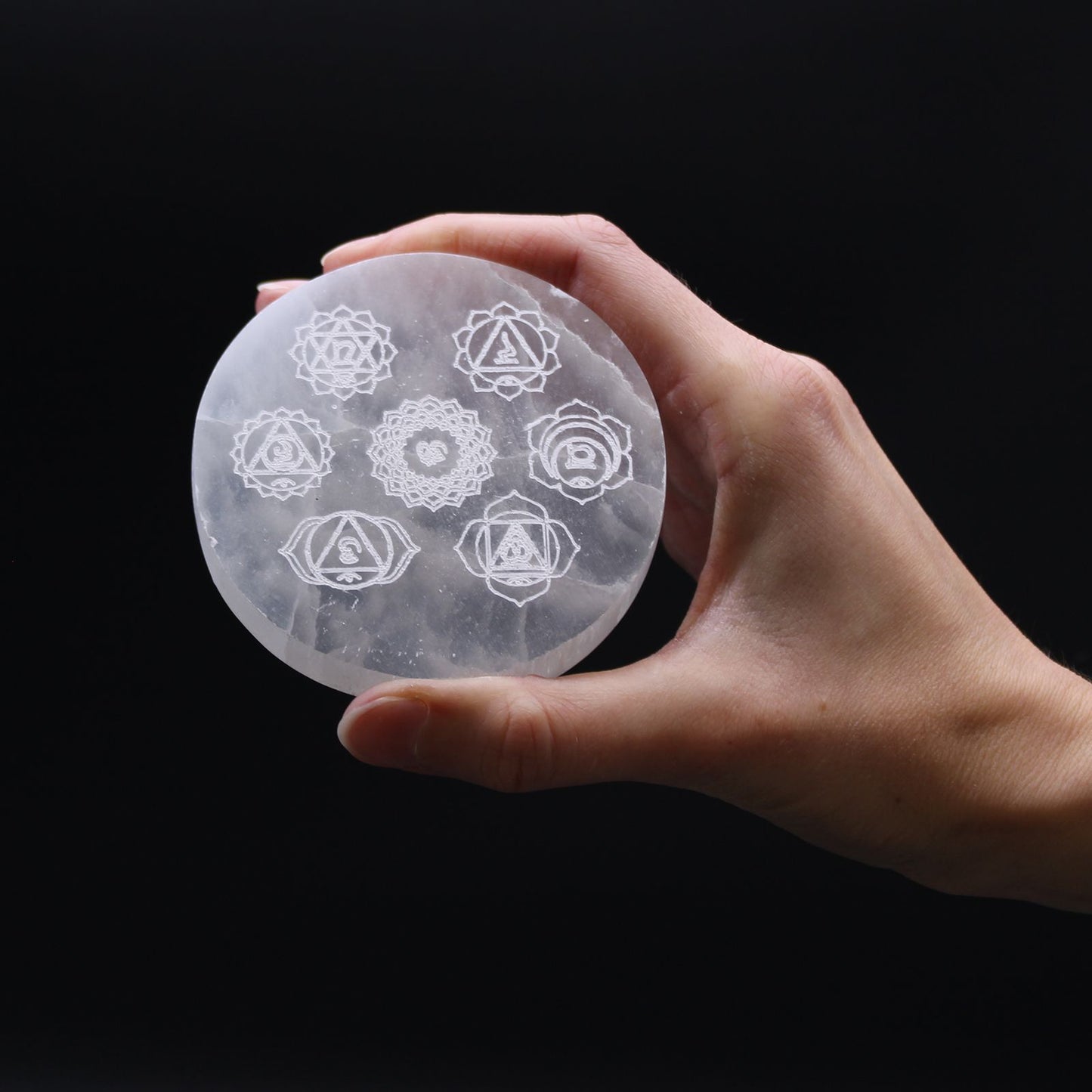 Round white selenite charging plate engraved with seven colourful chakra symbols on a wooden surface.