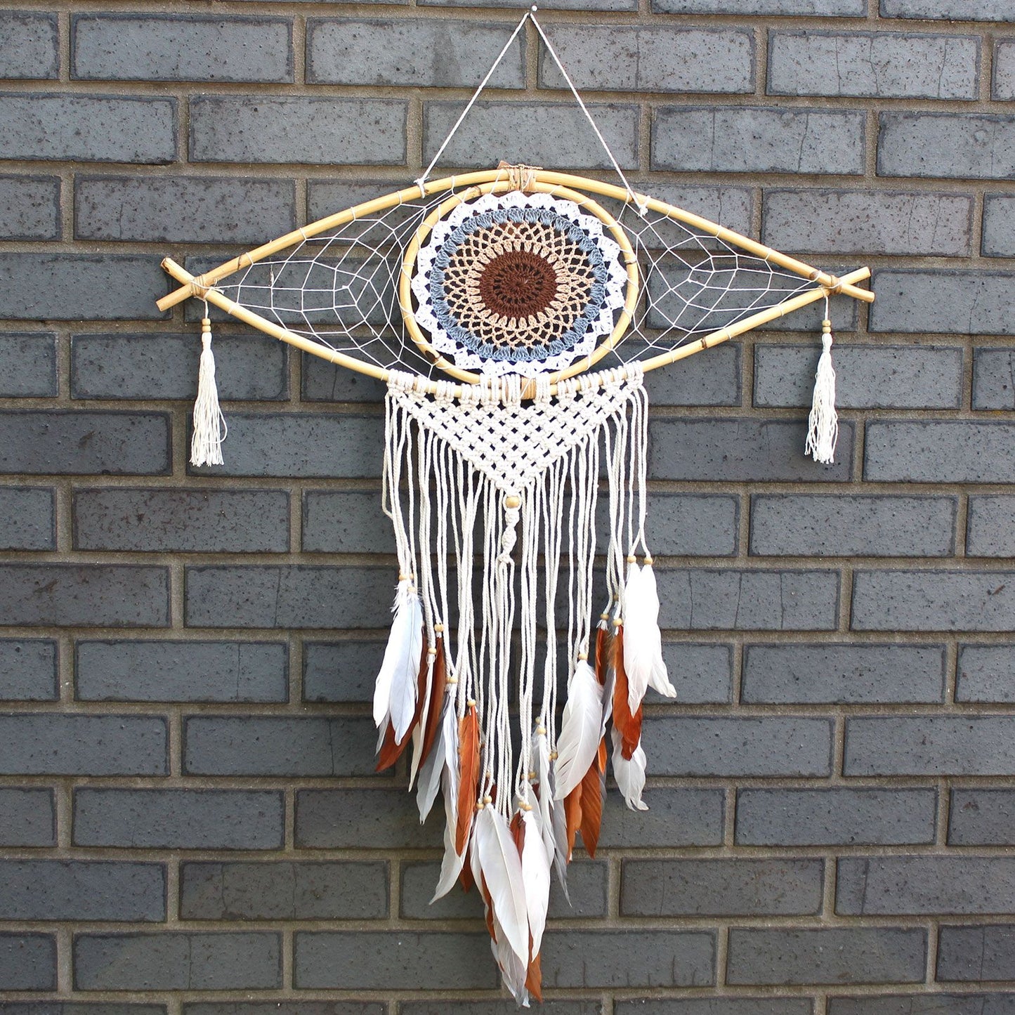 Large white, grey, and brown macrame dream catcher with a central evil eye design and hanging woven tails.