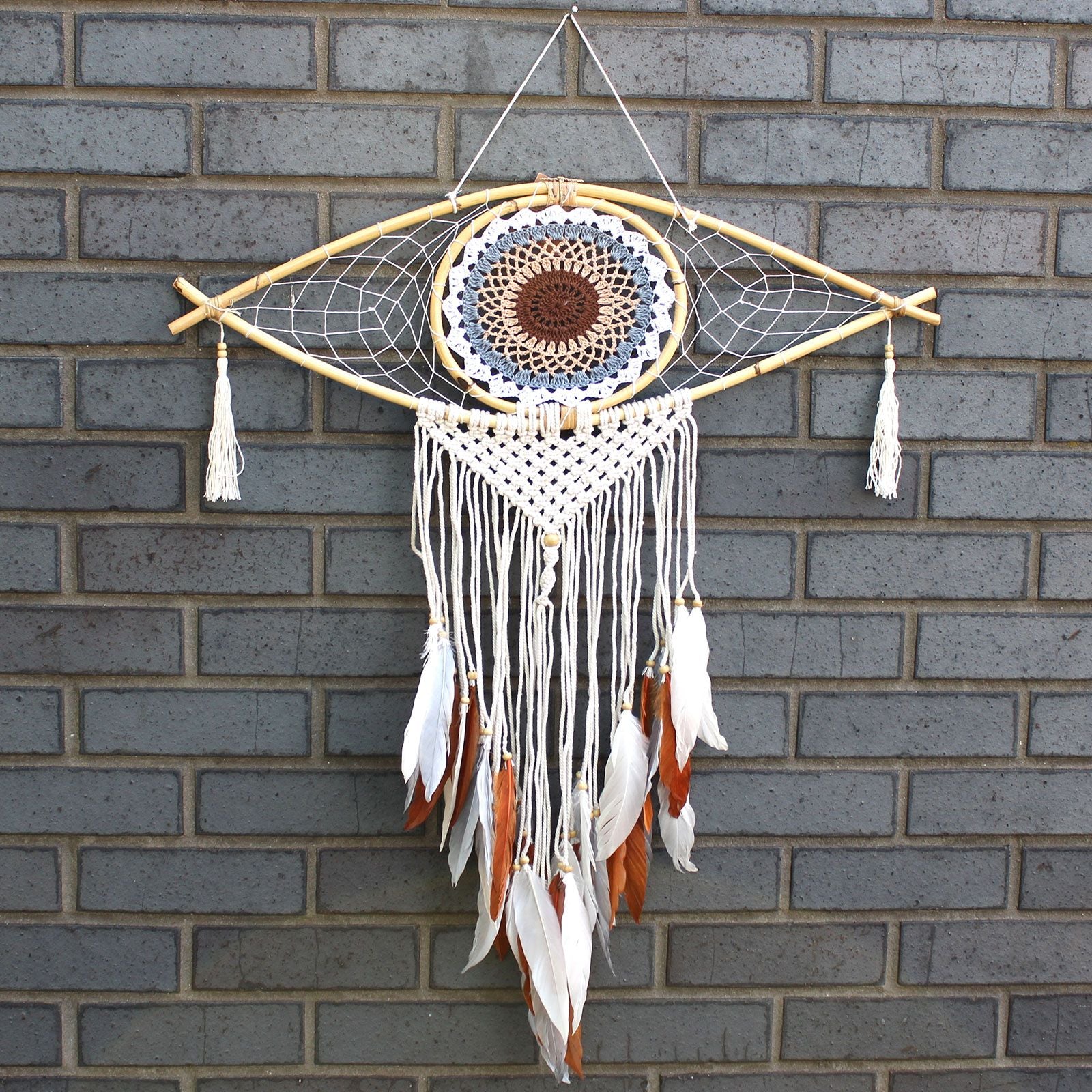 Large white, grey, and brown macrame dream catcher with a central evil eye design and hanging woven tails.