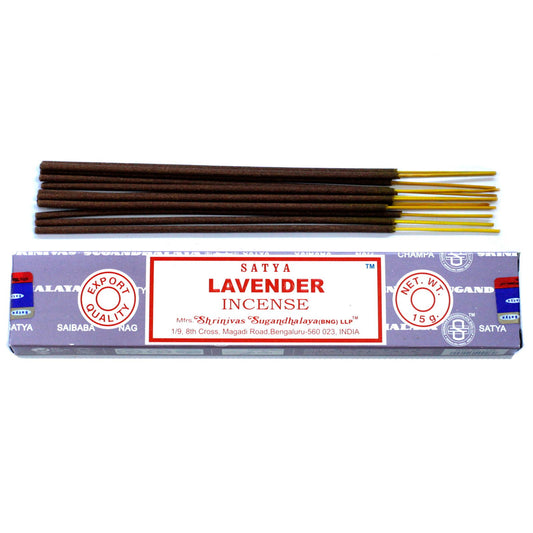 A pack of hand-rolled lavender incense sticks beside a small pile of purple lavender flowers on a neutral surface.