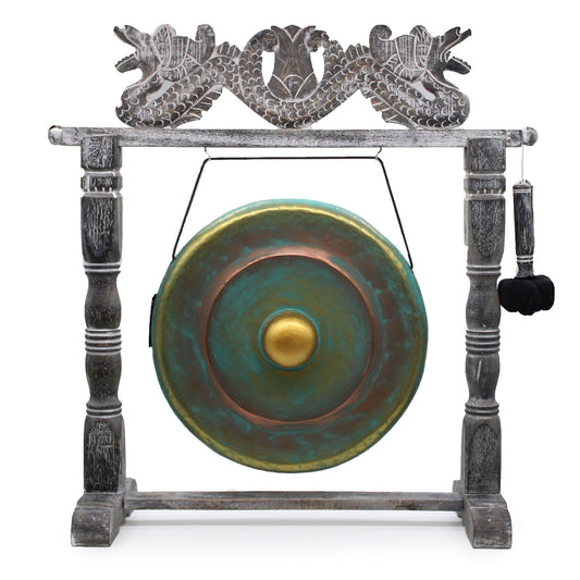 Handcrafted circular metal gong with a greenwash finish suspended in an ornate, dark wood-carved standing frame.