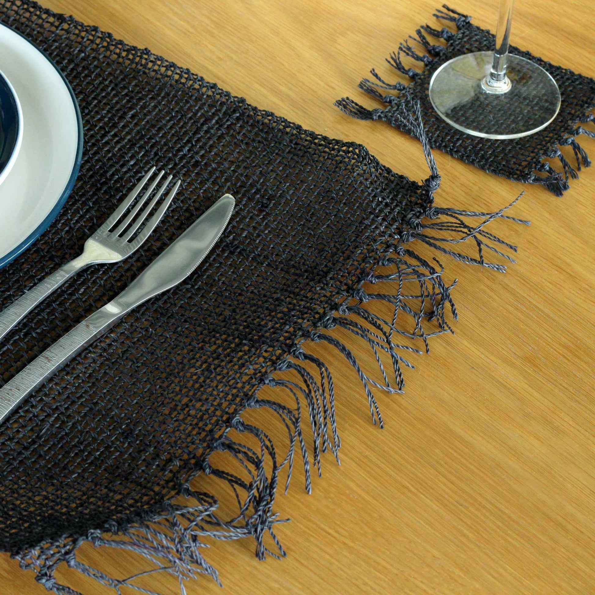 Round natural seagrass placemat with a bold black finish and decorative fringe detailing on a minimalist table setting.