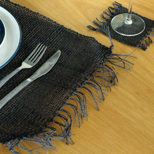 Round natural seagrass placemat with a bold black finish and decorative fringe detailing on a minimalist table setting.